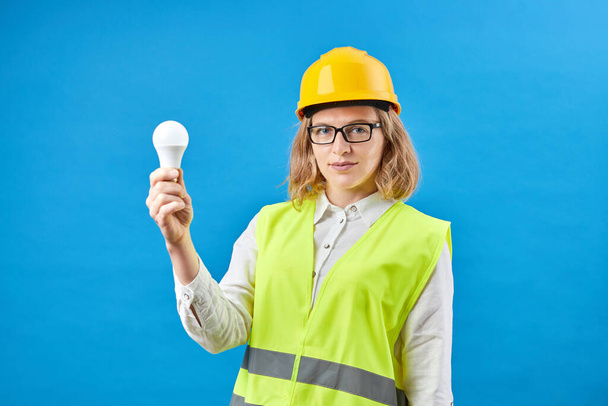 Junge Ingenieurin mit Arbeitsweste und gelbem Helm auf dem Kopf hält Glühbirne, während sie im Studio auf blauem Hintergrund steht - Foto, Bild