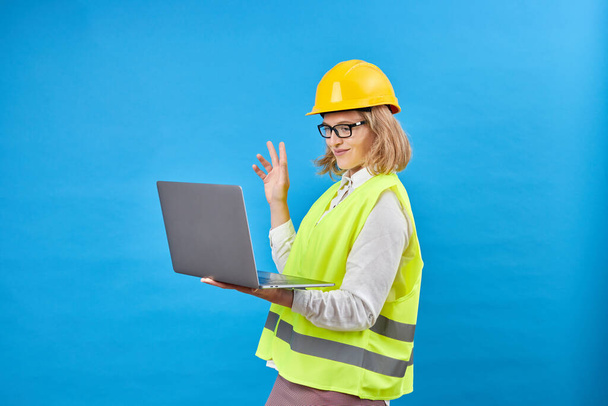 Studio shot of female professional engineering foreman manager wear hard helmet and reflective safety vest standing and smiling, holding laptop computer in studio on blue background - Foto, afbeelding