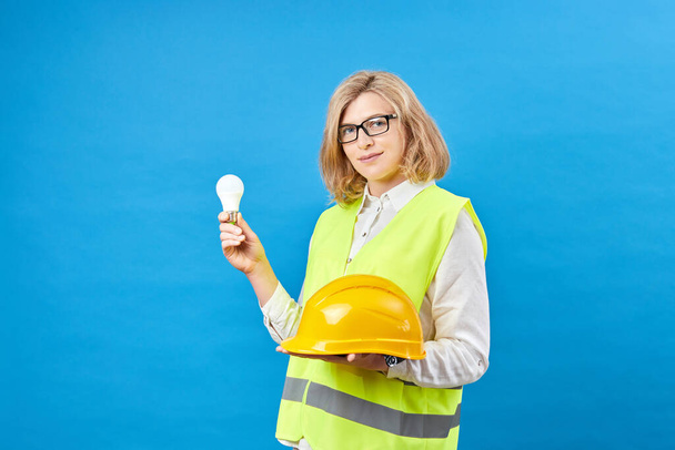 Young female engineer wearing work clothes holding a yellow helmet in one hand and holding a light bulb in the other hand while standing in a studio on a blue background - Fotoğraf, Görsel
