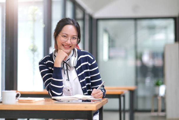 Asian woman wearing headphones using laptop in cafe, writing notes, attractive female student learning language, watching online webinar, listening audio course, e-learning education concept - 写真・画像