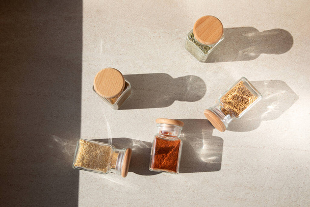 A group of seasoning in glass jars on a light stone background with shadows. Paprika, herbs, mustard, garlic, flat lay - Foto, Bild