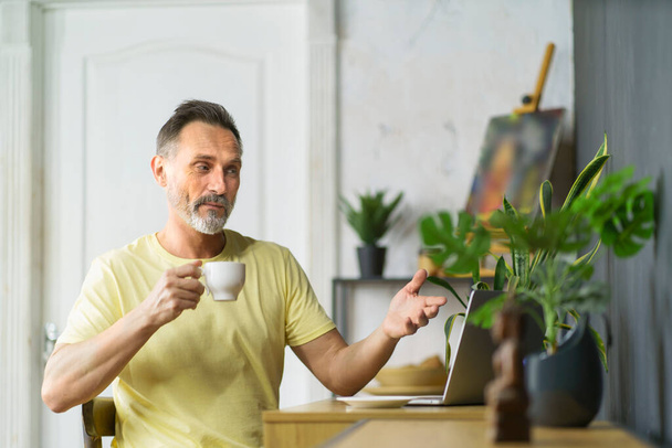 Bearded man looking at laptop while working at home office with video call or meeting holding cup of coffee. Middle age man gives video consulting on business issues from home office - Foto, Imagen