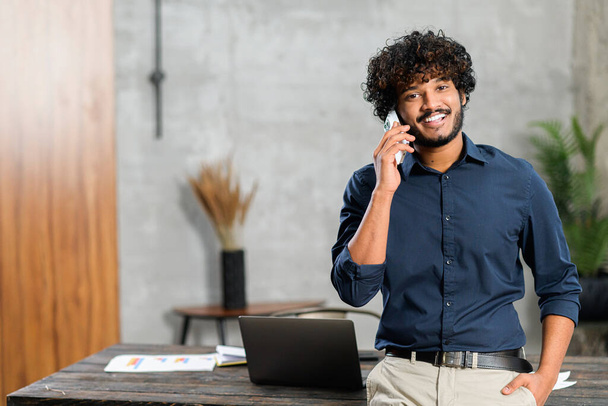 Happy inspired Indian male small business owner talking on the smartphone while standing in modern loft office. Smiling successful contemporary man manager in casual wear holding phone call,copy space - Photo, Image