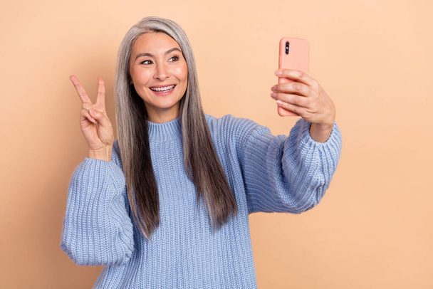 Photo of charming grey hair mature woman in comfy knitted sweater take selfie on phone show v-sign isolated on beige color background. - Photo, image