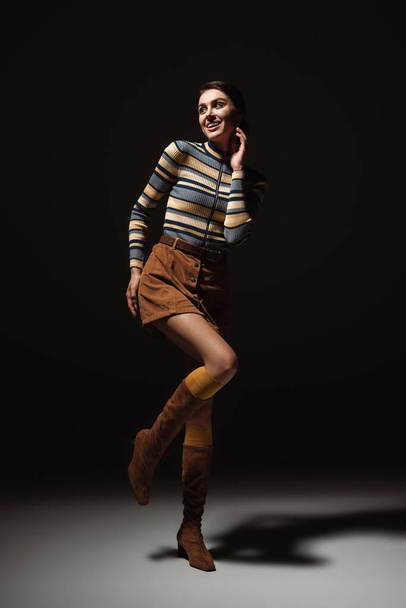 full length of cheerful young woman in turtleneck with skirt and boots posing on black  - Foto, imagen