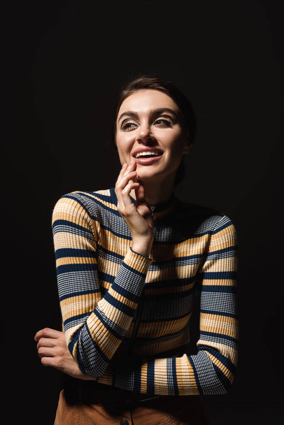 happy young woman in striped turtleneck looking away isolated on black  - Fotó, kép