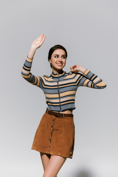 Ευτυχισμένη νεαρή γυναίκα με ζιβάγκο και φούστα χειρονομώντας ενώ ποζάρει απομονωμένη σε γκρι - Φωτογραφία, εικόνα