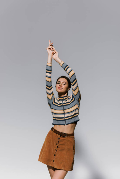 happy young woman in striped turtleneck and skirt posing with raised hands isolated on grey - Fotó, kép