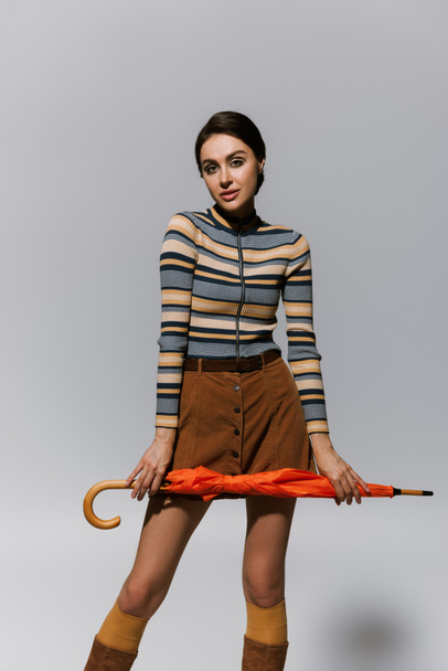brunette young woman in turtleneck and skirt posing with orange umbrella isolated on grey  - Foto, imagen