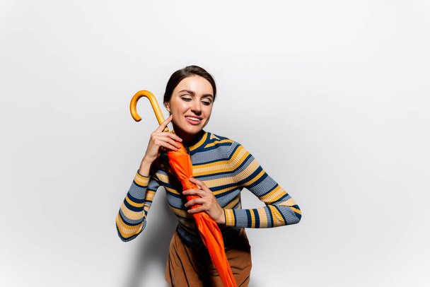 jovem mulher positiva em gola alta listrada posando com guarda-chuva laranja no branco - Foto, Imagem