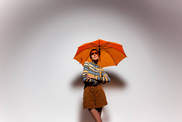 brunette young woman in striped turtleneck and skirt standing under orange umbrella on grey - Photo, Image