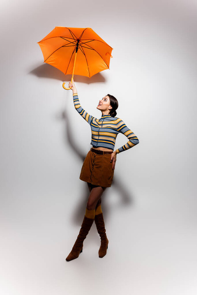 full length of cheerful woman in striped turtleneck and skirt posing with umbrella on grey - Fotografie, Obrázek