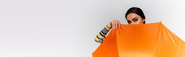 morena mujer joven cubriendo la cara con paraguas naranja aislado en gris, pancarta - Foto, Imagen
