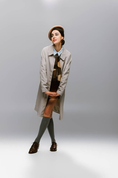 full length of elegant young woman in beret and trench coat looking away on grey - Foto, Imagem