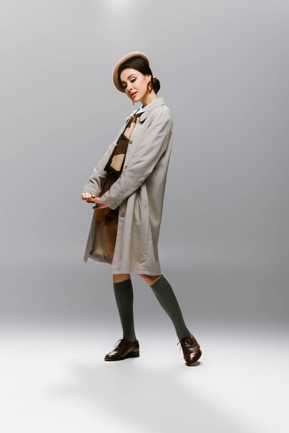 full length of elegant young woman in beret and stylish trench coat posing on grey - Foto, imagen