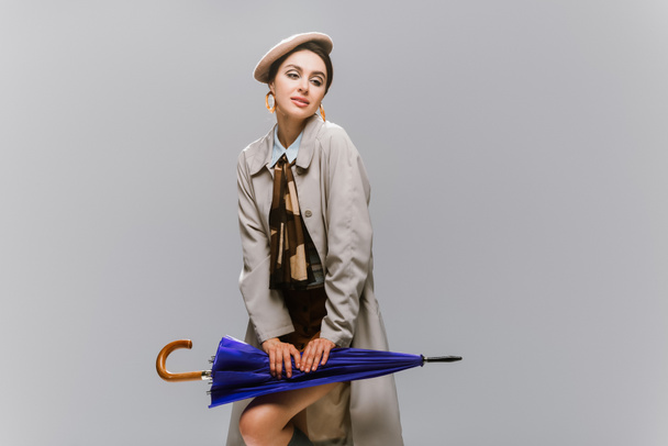 elegant young woman in beret and trench coat posing with umbrella isolated on grey - Foto, afbeelding