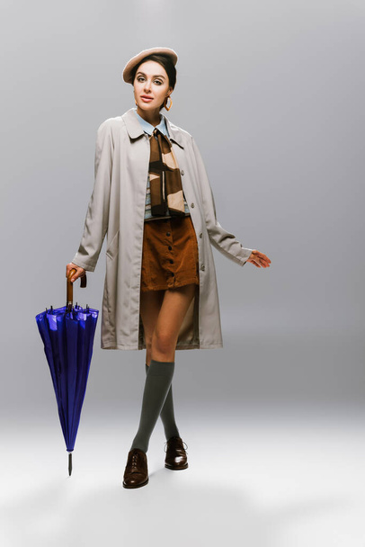 full length of brunette young woman in beret and trench coat posing with blue umbrella on grey - Foto, immagini