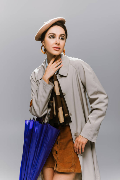 young woman in beret and trench coat posing with blue umbrella isolated on grey - Photo, Image
