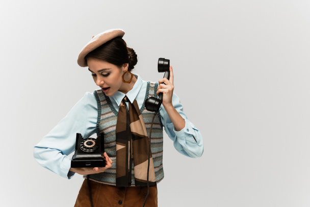 mujer joven asombrada en boina sosteniendo teléfono vintage aislado en gris  - Foto, Imagen