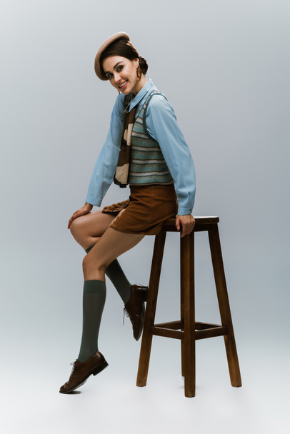 full length of happy young woman in beret and autumnal clothes sitting on wooden high chair on grey - Photo, Image