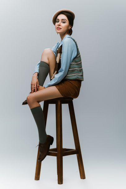 full length of young woman in beret and autumnal clothes sitting on wooden high chair on grey - Zdjęcie, obraz