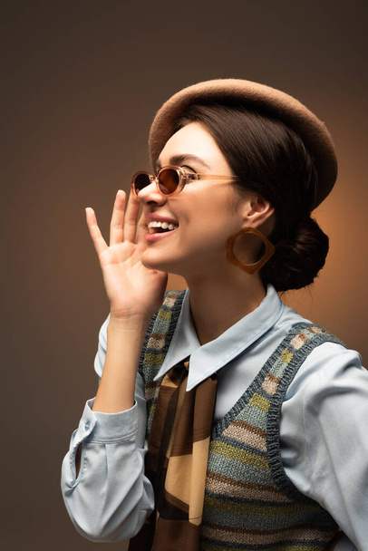 happy young woman in beret adjusting stylish sunglasses on brown  - Фото, зображення