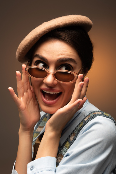 excited young woman in beret adjusting stylish sunglasses on brown  - Foto, afbeelding