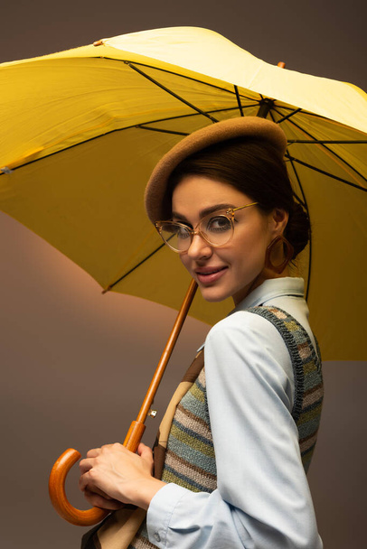 χαρούμενη νεαρή γυναίκα με μπερέ και γυαλιά που κρατά κίτρινη ομπρέλα στο γκρι - Φωτογραφία, εικόνα