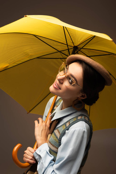 mujer joven positiva en boina y gafas con paraguas amarillo sobre gris - Foto, imagen