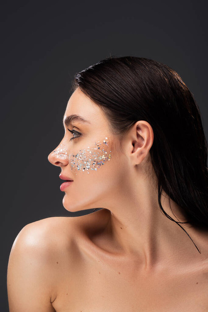 side view of pretty woman with bare shoulder and sparkling glitter on cheeks isolated on grey - Fotó, kép