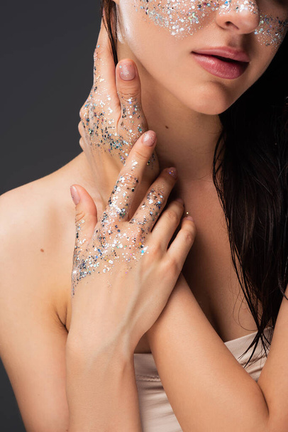 cropped view of young brunette woman with sparkling glitter on cheeks and hands touching neck isolated on grey  - 写真・画像
