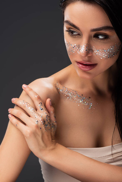 portrait of woman with natural makeup and sparkling glitter on face and body touching bare shoulder isolated on grey - Фото, изображение