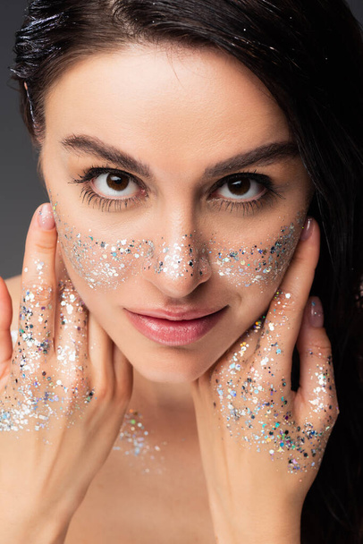 portrait of woman with natural makeup and glitter on cheeks and hands touching face while looking at camera isolated on grey - Фото, изображение