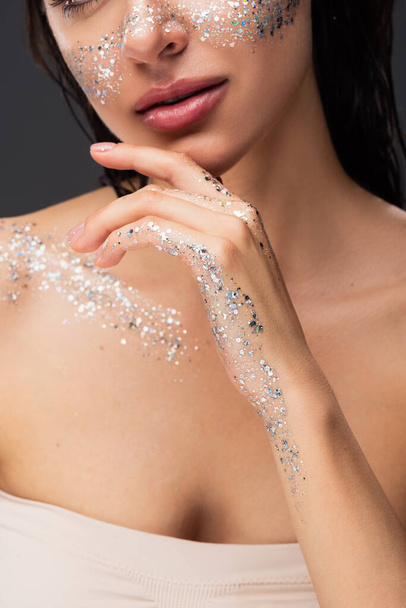 cropped view of young woman with sparkling glitter on cheeks and body touching chin isolated on grey  - Photo, Image