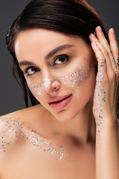 young woman with sparkling glitter on cheeks and body adjusting hair ad looking at camera isolated on grey  - Foto, Imagen
