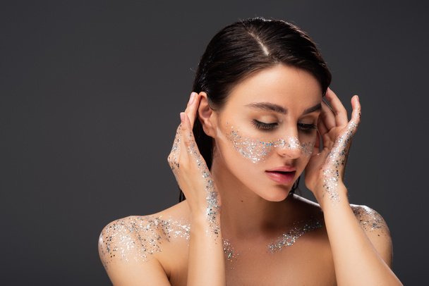 portrait of young woman with glitter on cheeks and hands adjusting hair isolated on grey  - Photo, Image