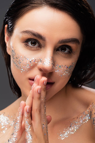 portrait of young woman with glitter on cheeks and praying hands looking at camera isolated on grey  - Foto, immagini