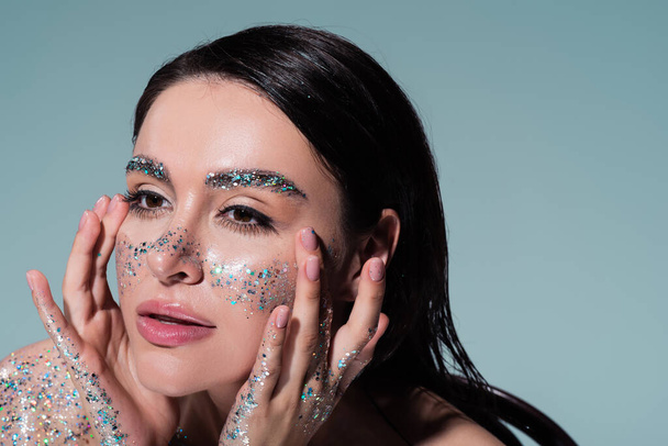 portrait of brunette woman with glitter on cheeks and eyebrows touching face while looking away isolated on grey  - Photo, Image