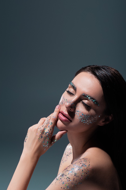 portrait of young brunette woman with closed eyes and glitter on eyebrows touching face isolated on grey  - Photo, Image