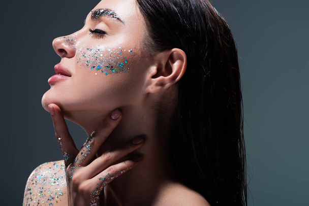 portrait of young brunette woman with closed eyes and glitter on eyebrows touching neck isolated on grey  - Foto, Imagen
