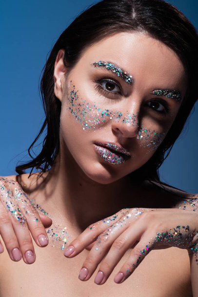 portrait of brunette young woman with glitter on lips and cheeks looking at camera isolated on blue  - 写真・画像