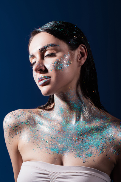 portrait of brunette young woman with closed eyes and glitter on body isolated on blue  - Foto, Imagem