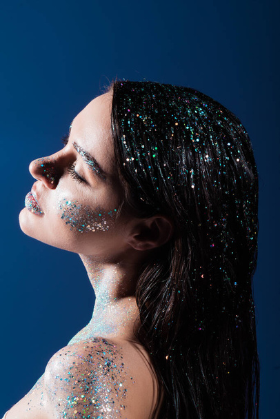 Seitenansicht der brünetten jungen Frau mit funkelndem Glitzern auf Körper und Haar isoliert auf blau  - Foto, Bild