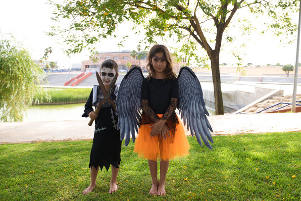 Ένα κορίτσι ντυμένο μάγισσα με πορτοκαλί φούστα και μαύρα φτερά και ένα αγόρι ντυμένο ζόμπι τρομάζουν την κάμερα σε ένα αποκριάτικο πάρτι. Φάρσα ή κέρασμα. 31 Οκτωβρίου. Καλό Χάλογουιν. - Φωτογραφία, εικόνα