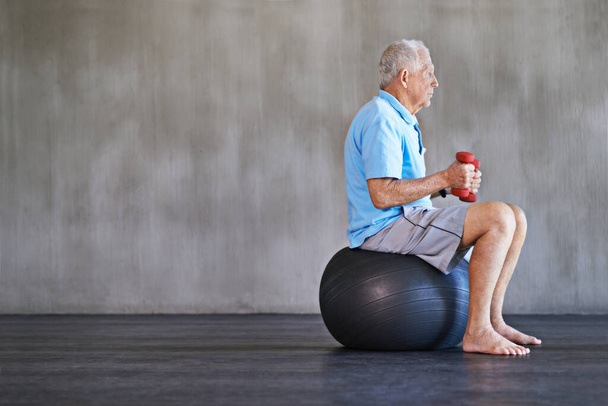 Yaşlanmaya karşı savaşmak. Yaşlı bir adam İsviçre topunun üzerinde otururken ağırlık kullanıyor. - Fotoğraf, Görsel