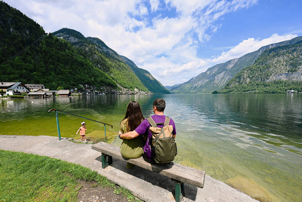 Couple with baby girl sitting on bench over Austrian alps lake in Hallstatt, Salzkammergut, Austria. - Foto, afbeelding