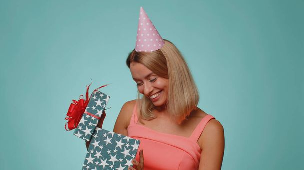 Cheerful young woman opening gift box, unwrapping birthday surprise and expressing great happiness amazement, satisfied with best present bonus. Pretty girl isolated alone on studio blue background - Photo, image