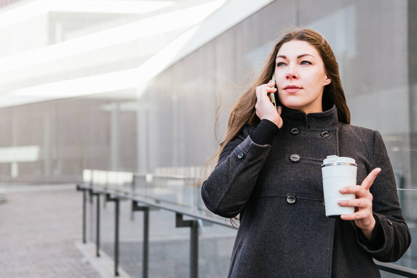 Ocupado fêmea em casaco quente de pé com copo térmica tumbler falando no celular em dia ventoso frio - Foto, Imagem