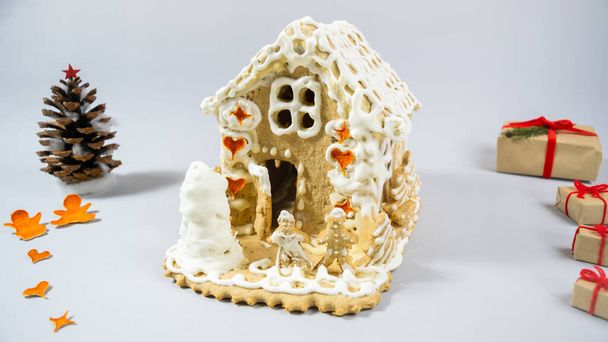 Homemade gingerbread house painted with white sweet icing and decorated with a cone, dried tangerine peels and gift boxes. Gray background. Concept of preparing delicious handmade sweets for holiday - Фото, изображение