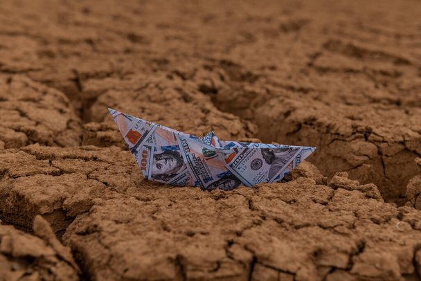 Papierowa łódź z wizerunkiem banknotu 100 dolarowego stoi na popękanej suchej gliny na pustyni - Zdjęcie, obraz